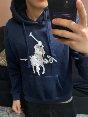 Mörkblå hoodie från Ralph Lauren - Snygg mörkblå hoodie från Ralph Lauren med stor vit logga och polospelare på bröstet. Klassisk känguruficka framtill och justerbar huva med dragsko. Perfekt för en avslappnad och trendig stil. Tillverkad i mjukt bomullsmaterial.