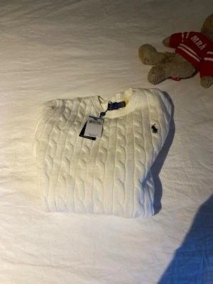 Vit kabelstickad tröja Ralph Lauren - Kabelstickad vit tröja från Ralph Lauren med rund halsringning och den klassiska broderade loggan på bröstet. Tröjan har långa ärmar och ribbade muddar vid ärmslut och nederkant. Perfekt för dig som gillar stilrena och tidlösa plagg. Obs rep är storleken S men passar även XS. Billigare vid snabb affär 