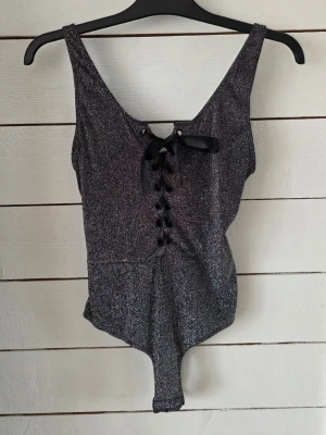 Glittrig svart body från Bik Bok - Snygg svart body från Bik Bok Lingerie med glittrigt tyg och snörning framtill. Djup ringning och dekorativ rosett ger en trendig look. Stretchigt material som sitter bekvämt och formar sig efter kroppen. Perfekt för dig som vill ha något extra till outfiten.