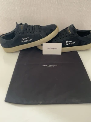 Svarta Saint Laurent sneakers - Svarta sneakers från Saint Laurent med vit text på sidan och beige sula. Skorna har klassisk låg profil, snörning och är tillverkade i mocka. Guldiga detaljer på plösen ger en exklusiv touch. Perfekta för dig som gillar stilrena och trendiga skor.