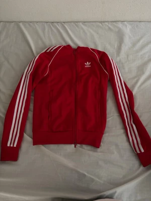 Röd Adidas zip hoodie med vita ränder - Snygg röd hoodie från Adidas med dragkedja och de klassiska tre vita ränderna längs ärmarna. Adidas-logga på bröstet och ribbade muddar. Tillverkad i mjukt material, perfekt för en sportig och avslappnad stil.