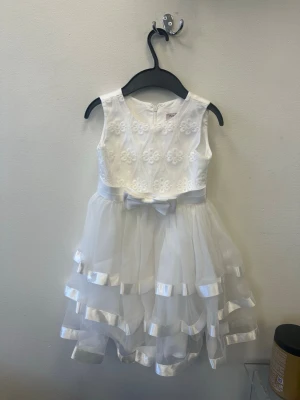 Vit ärmlös klänning med rosett - Söt vit ärmlös klänning med broderade blommor på överdelen och en fin rosett i midjan. Kjolen är i flera lager tyll med satinkanter för extra volym och en festlig look. Perfekt för speciella tillfällen.