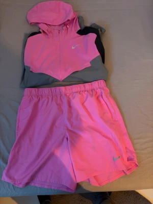 Nike rosa träningsset med hoodie - Snyggt träningsset från Nike med en rosa och grå hoodie med dragkedja och huva samt matchande rosa träningsshorts. Båda plaggen har Nike-logga och är tillverkade i lätt, ventilerande material som passar perfekt för löpning eller gym. Koftan är i storlek S och shortsen i Storlek L.