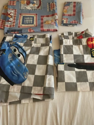 Bäddset Cars och nallemotiv - Säljer ett bäddset med motiv  på nallar-örngott och påslakan .Cars gardiner längder har stora tryck av bilar i blått och rött. Nallesetet har blå botten med rutiga detaljer och söta nallar. Perfekt för barnrummet och för dig som gillar färgglada motiv.