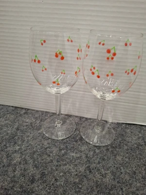 Två vinglas med körsbärsmotiv - Säljer två genomskinliga vinglas i glas med sött körsbärsmönster i rött och grönt. Perfekta för att piffa upp dukningen med en lekfull touch. Glasen har klassisk form med rund kupa och smal fot.