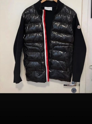 Moncler cardigan - Snygg svart dunväst från Moncler med glansig yta och quiltad design. Västen har stickade svarta ärmar, rödvit dragkedja och Moncler-logga på ärmen. Perfekt för lager-på-lager och streetstyle. Materialet är en mix av dun och stickat tyg.
