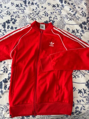 Röd Adidas track jacket med vita ränder - Snygg röd Adidas track jacket med dragkedja och klassiska vita ränder längs ärmarna. Tröjan har ståkrage, två fickor och Adidas logga broderad på bröstet. Tillverkad i polyester, perfekt för en sportig och avslappnad stil.