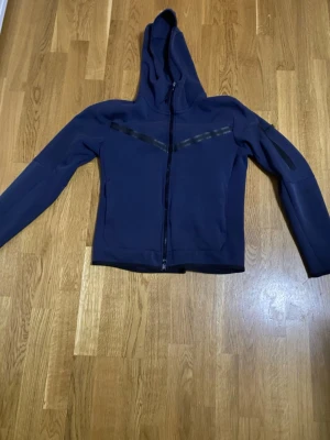 Mörkblå hoodie med dragkedja - Snygg mörkblå hoodie med hel dragkedja framtill och huva. Har diskreta svarta detaljer över bröstet och en ficka med dragkedja på ena ärmen. Tillverkad i ett mjukt och stretchigt material som känns sportigt och modernt.
