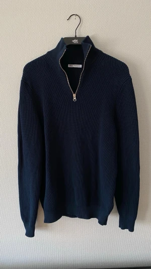 Quarterzip Zara - Snygg mörkblå stickad tröja från Zara med halvzip och hög krage. Tröjan har långa ärmar och ribbstickad struktur som ger en clean och modern look. Perfekt till jeans eller chinos för en avslappnad stil.
