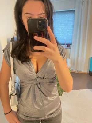 Glittrig silver topp med v-ringning - Snygg glittrig silver topp med v-ringning och twistad detalj framtill. Kortärmad modell i figurnära passform, perfekt för att sticka ut. Materialet är glansigt och stretchigt för en bekväm känsla.