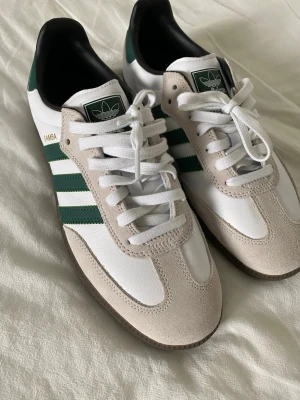 Adidas Samba vita och gröna sneakers - Klassiska Adidas Samba sneakers i vitt och grönt med beige mockadetaljer och bruna gummisulor. Skorna har de ikoniska tre ränderna på sidan och Adidas-logga på plösen. Ovandelen är i läder och mocka, och de har vita skosnören.