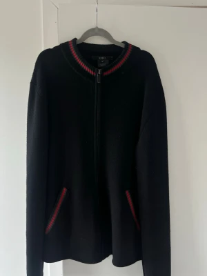 Svart ziptröja från Gucci - Svart stickad ziptröja från Gucci med ribbad krage och fickor, båda med rödgröna detaljer. Tröjan har en rak passform och långa ärmar. Perfekt för dig som gillar klassisk design med exklusiv känsla.