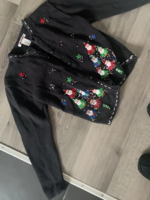 Vintage svart julkofta med tomtar från Air Pop Girl - Unik svart kofta från Air Pop Girl med färgglada broderade tomtar, julgranar och stjärnor framtill. Koftan har rund hals, knäppning fram och vita detaljer längs kanterna. Materialet är 55% ramie och 45% bomull. Perfekt för dig som vill sticka ut under julen!