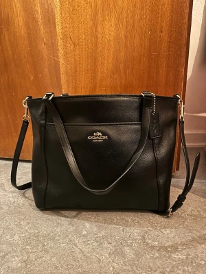 Svart handväska från Coach - Coach Crossgrain Leather Pocket Tote, modell F34497. Svart handväska från Coach i slätt läder med guldfärgade detaljer. Väskan har både axelrem och handtag, samt en praktisk ytterficka framtill med Coach-logga i guld. Innerfodret i ytterfacket är trasigt annars är den i mycket bra skick. Lädret är behandlat och tål rimlig mängd vatten. Nypris 10 000 kr.