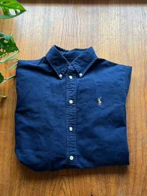 Mörkblå skjorta från Ralph Lauren - Säljer en mörkblå skjorta från Ralph Lauren med klassisk button-down krage och vita knappar. Skjortan har broderad logga på bröstet och är i bomull med custom fit-passning. Perfekt för dig som gillar stilrena plagg med ikonisk design.