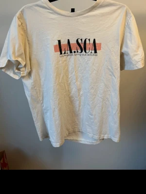 Beige t-shirt med LA.SCA tryck - Beige t-shirt med svart och rosa LA.SCA-tryck på bröstet och texten 'Los Angeles spring club activities' under. Klassisk rund hals och korta ärmar. Tillverkad i mjukt bomullstyg, perfekt för en avslappnad stil.