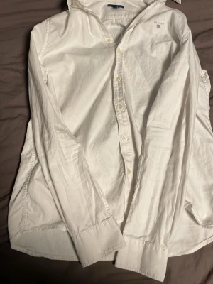 Vit klassisk skjorta från Gant - Klassisk vit skjorta från Gant med broderad logga på bröstet. Skjortan har lång ärm, knappar framtill och är tillverkad i mjuk bomull. Perfekt för en stilren och tidlös look.