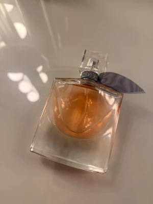 La Vie Est Belle parfym från Lancôme 50ml - La Vie Est Belle parfym från Lancôme i en elegant glasflaska med mjuka former och persikofärgad vätska. Flaskan har en genomskinlig fyrkantig kork och ett dekorativt grått band runt halsen. Perfekt för dig som gillar lyxiga och feminina dofter.