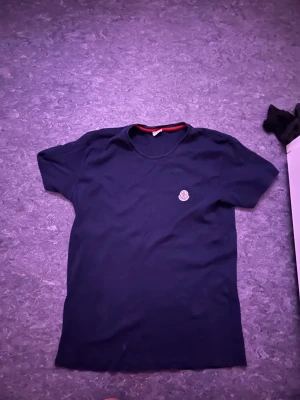 Mörkblå t-shirt från Moncler - Säljer en mörkblå t-shirt från Moncler med klassisk rund hals och korta ärmar. T-shirten har en liten Moncler-logga på bröstet och en röd detalj längs insidan av halsen. Perfekt för dig som gillar stilrena plagg med exklusiv känsla.