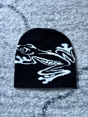 Svart mössa med vit groda  - Säljer en svart mössa med en stor vit grafisk illustration på framsidan. One size storlek och unisex. För vuxna. Mössan har en klassisk form och är stickad i mjukt material som passar perfekt för kyligare dagar. Perfekt accessoar för dig som vill sticka ut lite extra.