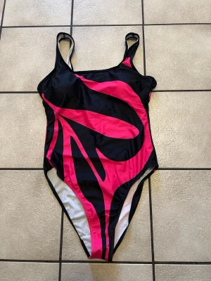 Svart och rosa baddräkt med mönster storlek L - Snygg baddräkt i svart med starkt rosa abstrakt mönster. Klassisk modell med smala axelband och hög benskärning. Materialet är troligtvis stretchigt syntetmaterial som polyester eller elastan, perfekt för simning eller strandhäng.