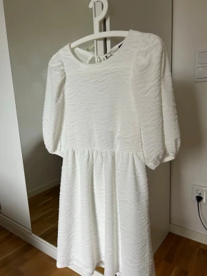 Vit kortklänning med snörning i ryggen - Supersöt vit kortklänning med strukturmönster och puffiga trekvartsärmar. Klänningen har öppen rygg med snygga snörningsdetaljer och rund halsringning. Perfekt för sommarens alla tillfällen!