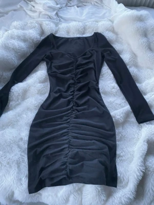 Svart långärmad klänning - Snygg svart klänning med långa ärmar och rynkade detaljer framtill. Klänningen har en kroppsnära passform och är tillverkad i ett stretchigt material som ger en smickrande siluett. Perfekt för dig som gillar stilrena och trendiga plagg.
