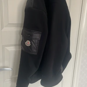 Moncler cardigan  - Snygg svart bomberjacka från Moncler med en ficka på ärmen och Moncler-logga. Jackan har ribbade muddar och är tillverkad i polyester med detaljer i skinn. Perfekt för dig som vill ha en stilren och trendig jacka. 