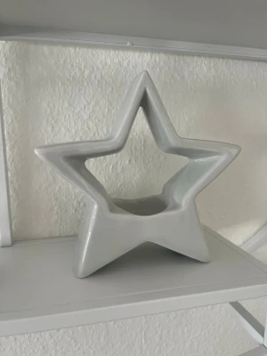 Vit stjärnformad ljushållare - Säljer en dekorativ ljushållare i form av en stjärna. Den är vit och tillverkad i keramik, perfekt som inredningsdetalj på hyllan eller bordet. Passar för värmeljus och ger ett stilrent intryck i rummet.