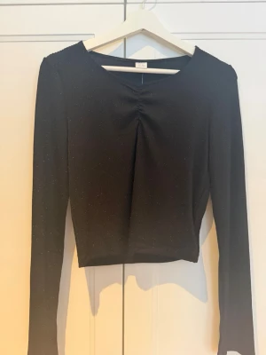 Svart glittrig långärmad topp - Snygg svart topp med lång ärm och diskret glittrigt tyg. Toppen har en lätt rynkning framtill vid halsen som ger en cool detalj. Knappt använd