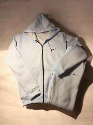 Ljusblå zip hoodie från Nike - Snygg ljusblå hoodie från Nike med dragkedja framtill och klassisk svart swoosh-logga på bröstet och ärmen. Hoodien har huva med dragsko och ribbade muddar vid ärmslut och nederkant. Perfekt för en avslappnad och sportig stil.