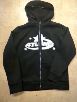 4TUNE Tröja - Svart hoodie med dragkedja och stor huva. På bröstet finns ett vitt tryck med texten '4TUNE' och stjärnor runtom. Hoodien har känguruficka framtill och ribbade muddar vid ärmar och nederkant. Perfekt för en avslappnad streetwear-look.