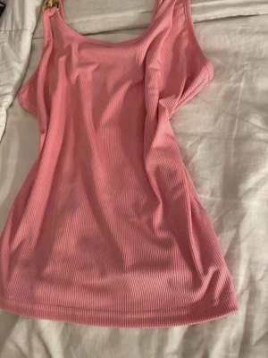 Rosa ribbad linnetopp med guldkedjor - Supersnygg rosa ribbad linnetopp med breda axelband dekorerade med stora guldfärgade kedjor. Toppen har en figurnära passform och är perfekt för att lyfta vilken outfit som helst. Materialet känns mjukt och stretchigt.