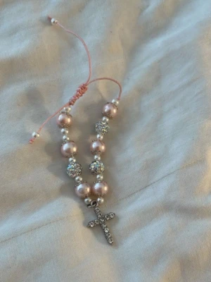 Rosa pärlarmband med korsberlock - Snyggt armband med rosa pärlor och glittriga stenar på elastisk tråd. Armbandet har en silverfärgad korsberlock med små strassdetaljer. Perfekt accessoar för dig som gillar bling och vill sticka ut lite extra.