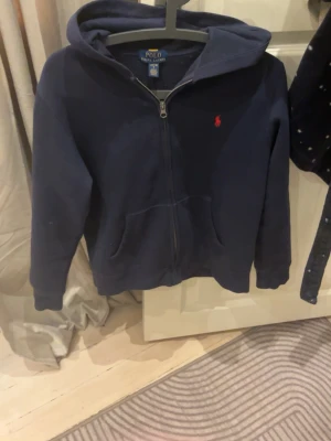 Mörkblå hoodie från Polo Ralph Lauren - Mörkblå hoodie med dragkedja från Polo Ralph Lauren. Klassisk design med huva, fickor framtill och den ikoniska röda logotypen broderad på bröstet. Perfekt för en avslappnad och stilren look. Tillverkad i mjukt material som känns skönt mot huden.