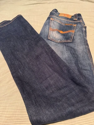Blå jeans från Nudie Jeans - Säljer ett par klassiska blå jeans från Nudie Jeans med snygg slitning och kontrastsömmar. Jeansen har en rak passform och den ikoniska bakfickan med orange brodyr. Tillverkade i slitstarkt denimtyg, perfekta för en avslappnad stil. W29L34. Hör av er om frågor eller mer bilder👍