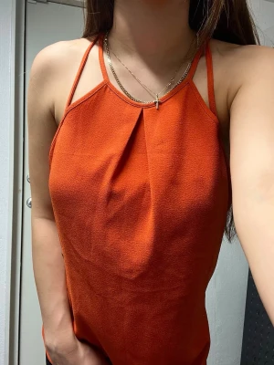 Orange linne med tunna axelband - Snyggt orange linne med tunna dubbla axelband och halterneck-skärning. Toppen har en lös passform och ett veck framtill som ger en cool detalj. Materialet är lätt och lite strukturerat, perfekt för varma dagar.
