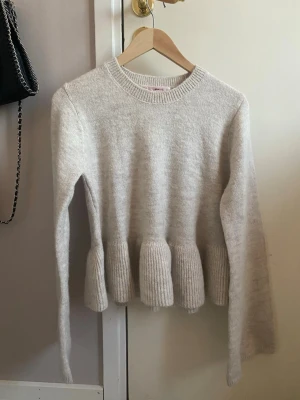 Beige stickad volangtröja - Supersöt beige stickad tröja med volangdetaljer nedtill. Tröjan har rund halsringning, långa ärmar och ribbstickade muddar. Perfekt för dig som gillar en feminin och trendig look. Materialet känns mjukt och skönt mot huden.