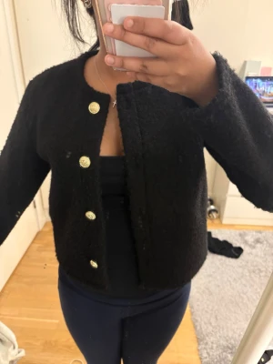 Svart kofta med guldfärgade knappar - Säljer en svart kofta med fyra guldfärgade knappar framtill. Koftan har en rund halsringning och är tillverkad i ett fluffigt material som ger en cool textur. Perfekt för att lyfta din outfit med en elegant touch.
