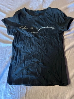 Svart t-shirt med texttryck - Svart t-shirt med vit text på bröstet där det står 'life is a journey, enjoy it'. Klassisk rund halsringning och korta ärmar. Tillverkad i mjuk bomull för en bekväm känsla. Perfekt till jeans eller shorts för en avslappnad look.