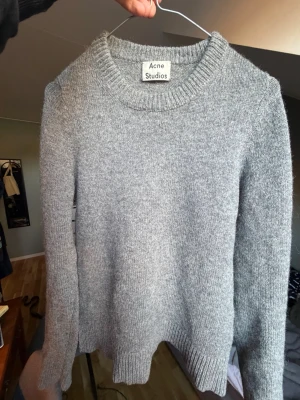 Grå stickad tröja från Acne Studios - Klassisk grå stickad tröja från Acne Studios med rund halsringning och ribbade muddar vid ärmslut och nederkant. Tröjan har en enkel och stilren design, perfekt för lager på lager och passar till det mesta. I 100 procent Shetlands Ull. Har två skavanker som är lagade som inte märks av. Använd Ca 20 gånger