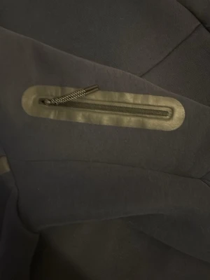 Mörkblå Nike tech  - Snygg mörkblå Nike Tech med dragkedja och luva. Tillverkad i ett mjukt och stretchigt material som känns bekvämt mot huden. Hoodien har en diskret ficka med dragkedja och stilrena sömmar för en modern look.