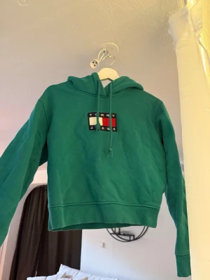 Grön hoodie från Tommy Jeans - Snygg grön hoodie från Tommy Jeans med klassisk logga broderad på bröstet i vitt, blått och rött. Hoodien har huva med dragsko och ribbade muddar vid ärmslut och nederkant. Perfekt för en avslappnad och trendig look.