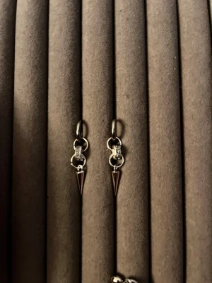 Silvriga örhängen med spetsdetalj - Säljer ett par silvriga örhängen med små ringar och en cool konformad spets som hänger ned. Örhängena har en minimalistisk och edgy design, perfekta för dig som gillar att sticka ut. Materialet ser ut att vara metall och de har en enkel klickstängning.