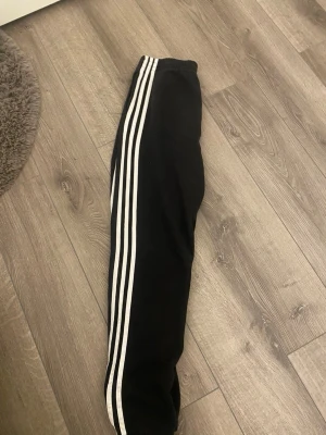 Svarta Adidas mjukisbyxor med ränder - Klassiska svarta mjukisbyxor från Adidas med tre vita ränder längs benen och elastisk midja. Byxorna har en avslappnad passform och är perfekta för chill eller träning. Adidas-logga på ena benet. Tillverkade i mjukt material för extra komfort.