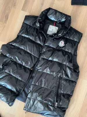 Svart dunväst från Moncler - Snygg svart dunväst från Moncler med glansig finish och hög krage. Västen har dragkedja framtill, två fickor med dragkedja och Moncler-logga på bröstet. Perfekt för lager-på-lager och streetstyle.
