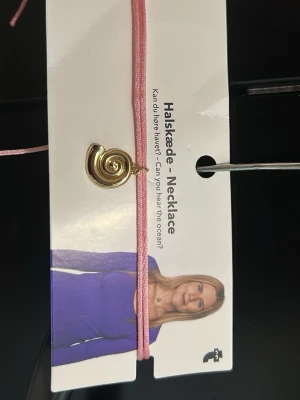 Rosa halsband med snäckhänge - Snyggt halsband med ett rosa band och ett guldigt snäckformat hänge. Perfekt accessoar för dig som gillar strandvibbar och unika detaljer. Halsbandet har en enkel men trendig design som passar till många olika stilar. Aldrig använd.