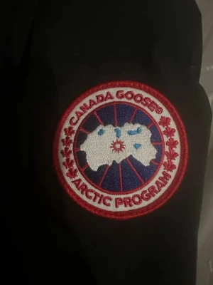 Svart dunjacka från Canada Goose - Säljer en svart dunjacka från Canada Goose med klassisk patch på ärmen och huva. Jackan har en stilren design med knappar och dragkedja framtill. Perfekt för kalla vinterdagar och riktigt varm tack vare dunfyllningen. Jag säljer den så snabbt möjligt 