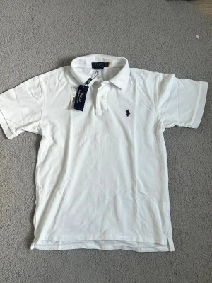 Vit pikétröja Polo Ralph Lauren - Klassisk vit pikétröja Polo Ralph Lauren med korta ärmar och krage. Tröjan har två knappar vid halsen och den ikoniska broderade loggan i marinblått på bröstet. Tillverkad i mjuk bomull som känns skön mot huden. Den är i storlek M men kan även passa S också. Jag har använt den 2-3 gånger och den är väldigt fin och bra.Fraktar samma dag eller nästa📦🚚 Aktiv hela tiden✅ hör av dig om du undrar något eller vill ha fler bilder⁉️