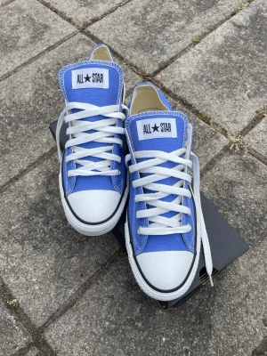 Blå Converse All Star sneakers - Snygga blå Converse All Star sneakers med klassisk låg modell. Skorna har vit tåhätta i gummi, vita snören och sula samt tydlig All Star-logga på plösen. Ovandelen är i canvas och sulan är i gummi. Perfekta för en avslappnad streetstil. De är helt oanvända och säljs du de var för stora. Orginal kartongen finns kvar. 